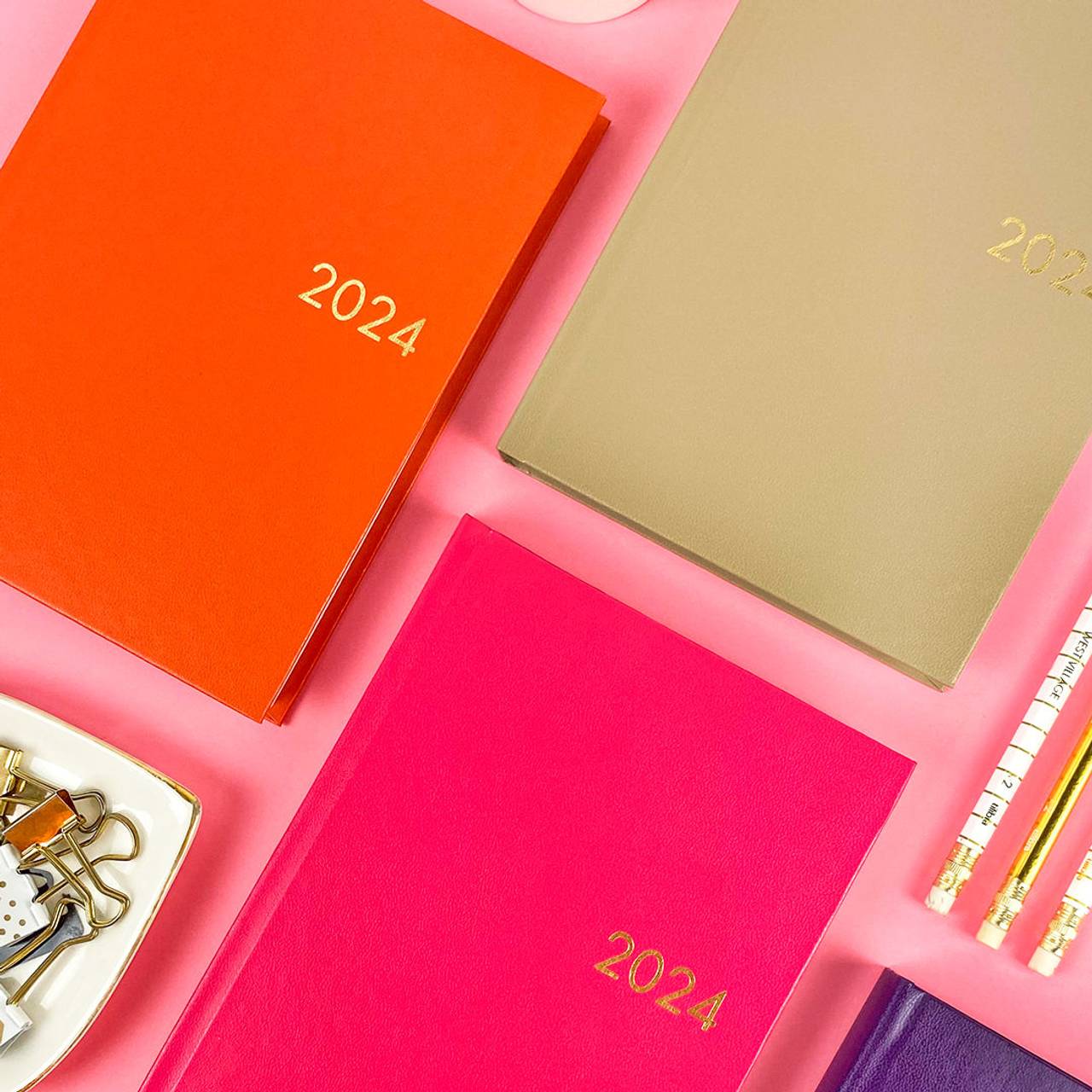Agendas Tilibra da coleção Napoli para 2024. Cores sólidas laranja, verde oliva e rosa com o ano em relevo dourado sobre fundo rosa claro.