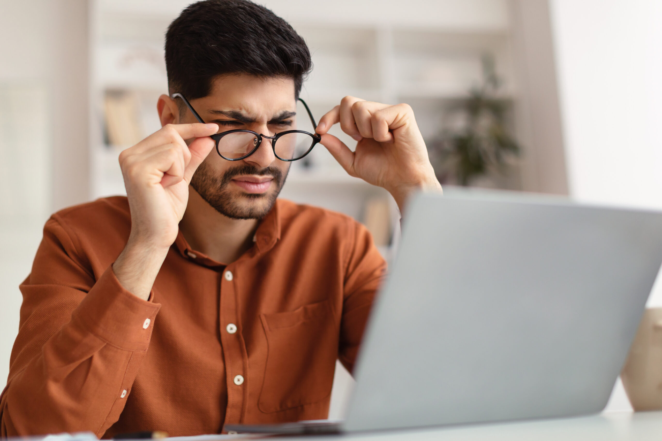 Homem afastando os óculos enquanto força os olhos tentando enxergar o conteúdo do computador a sua frente