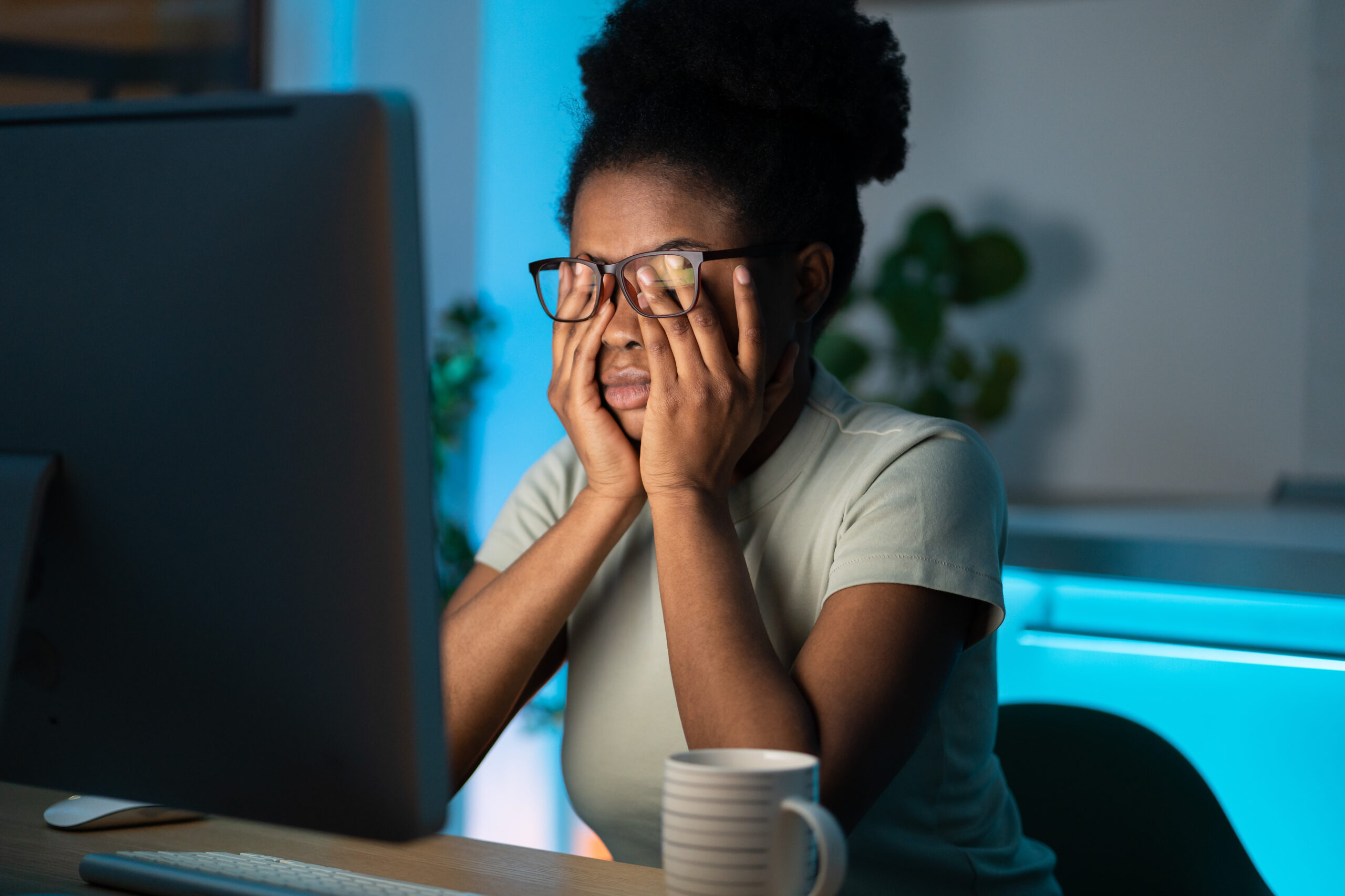 Mulher em frente ao computador com a mão sobre os olhos tocando por baixo dos óculos