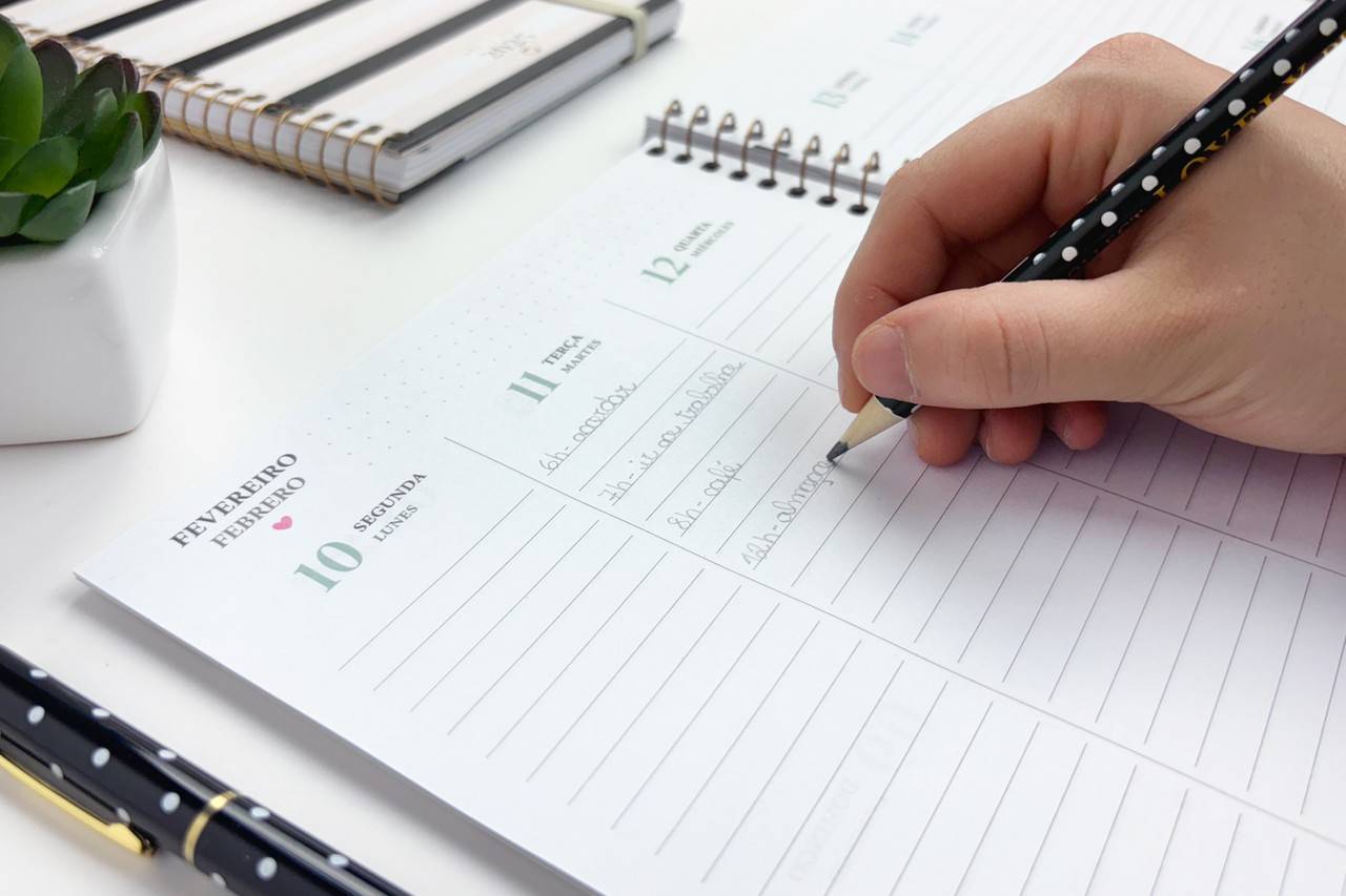 Mão segurando lápis escrevendo em uma agenda branca