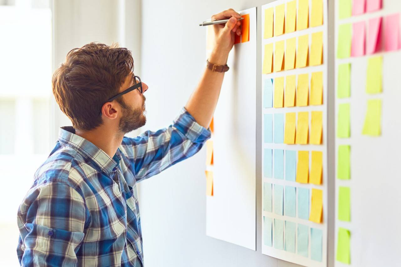 Homem fazendo anotações em um papel adesivo colodo em um quadro branco com diversos outros avisos colados ao redor