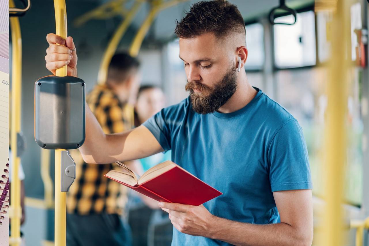 Homem em pé dentro de ônibus apoiado em barra vertical enquanto olha para baixo lendo um livro em sua outra mão