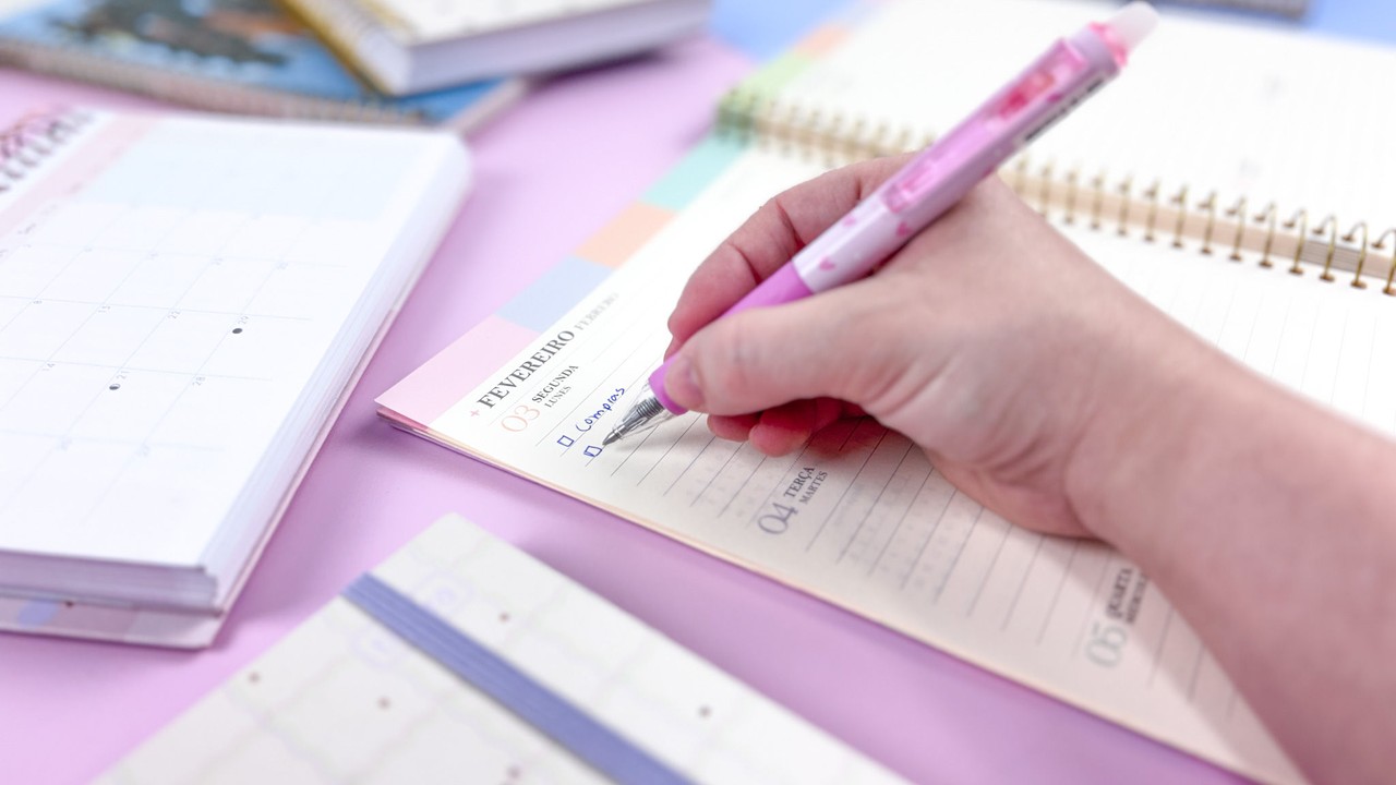 Mao de uma pessoa segurando uma caneta rosa escrevendo na folha de um caderno